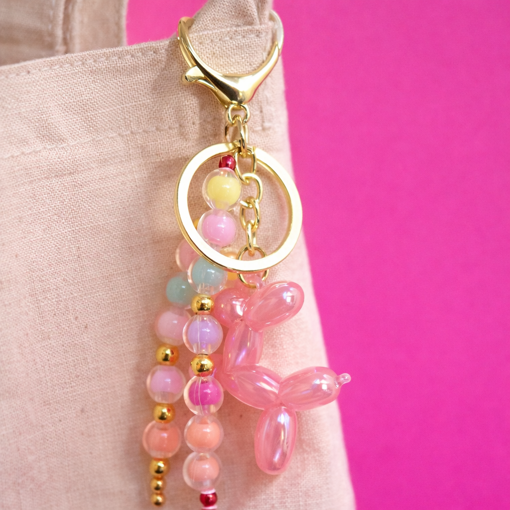 Detalle de un llavero con perrito de globo rosado y cuentas pastel, colgado de una totebag beige sobre fondo fucsia.