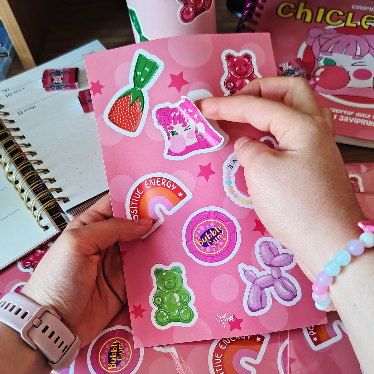 Manos despegan un sticker de una niña de cabello rosa de la lámina Chicle Rosa de Javipinkk, con dulces y ositos de goma.