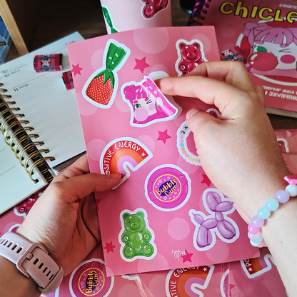 Manos despegan un sticker de una niña de cabello rosa de la lámina Chicle Rosa de Javipinkk, con dulces y ositos de goma.