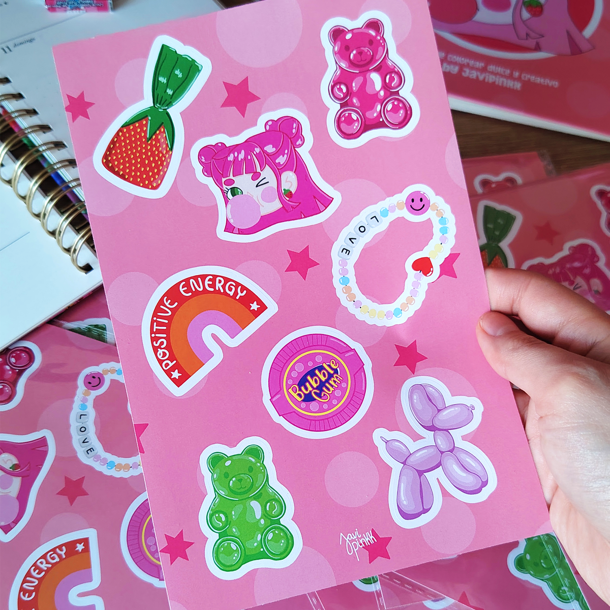 Mano sostiene una lámina de stickers Chicle Rosa de Javipinkk con ositos de goma, dulces, arcoíris y un perrito de globo.