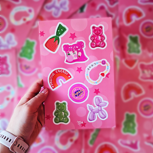 Mano sostiene una lámina de stickers Chicle Rosa de Javipinkk con ositos de goma, arcoíris, dulces y perrito de globo.