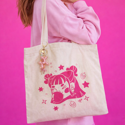 Persona con polerón rosa lleva una totebag beige Chicle Rosa de Javipinkk, con ilustración fucsia y colgante rosado.